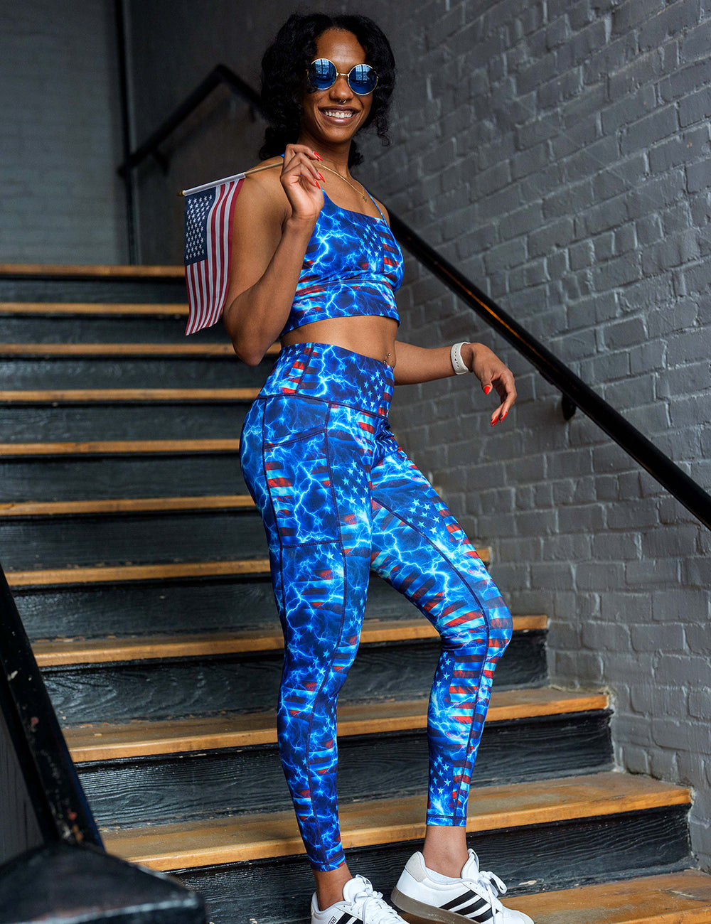 model wearing the lighting American flag patterned leggings and bra, holding a American flag and wearing blue sunglasses posing in a stairwell