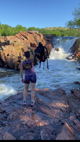 A person standing on rocky cliffs overlooking a powerful waterfall with a clear blue sky.