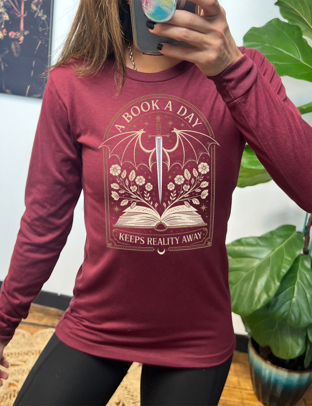 heather cardinal long sleeve shirt with the graphic of an archway, and within it, the graphic of an open book, with flowers growing out of it, the graphic of a sword poised downwards, with dragon wings, and many stars throughout, and the words saying "a book a day keeps reality away"