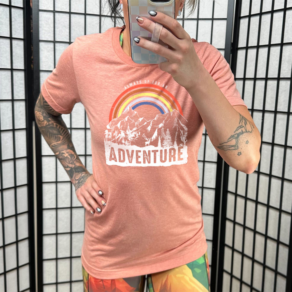 Model wearing sunset colored shirt with a rainbow over mountains graphic on it that says "Always Up For An Adventure"