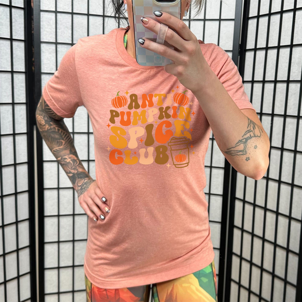 Model wearing sunset colored shirt with pumpkin and hot drink cup graphic on it that says "Anti Pumpkin Spice Club"