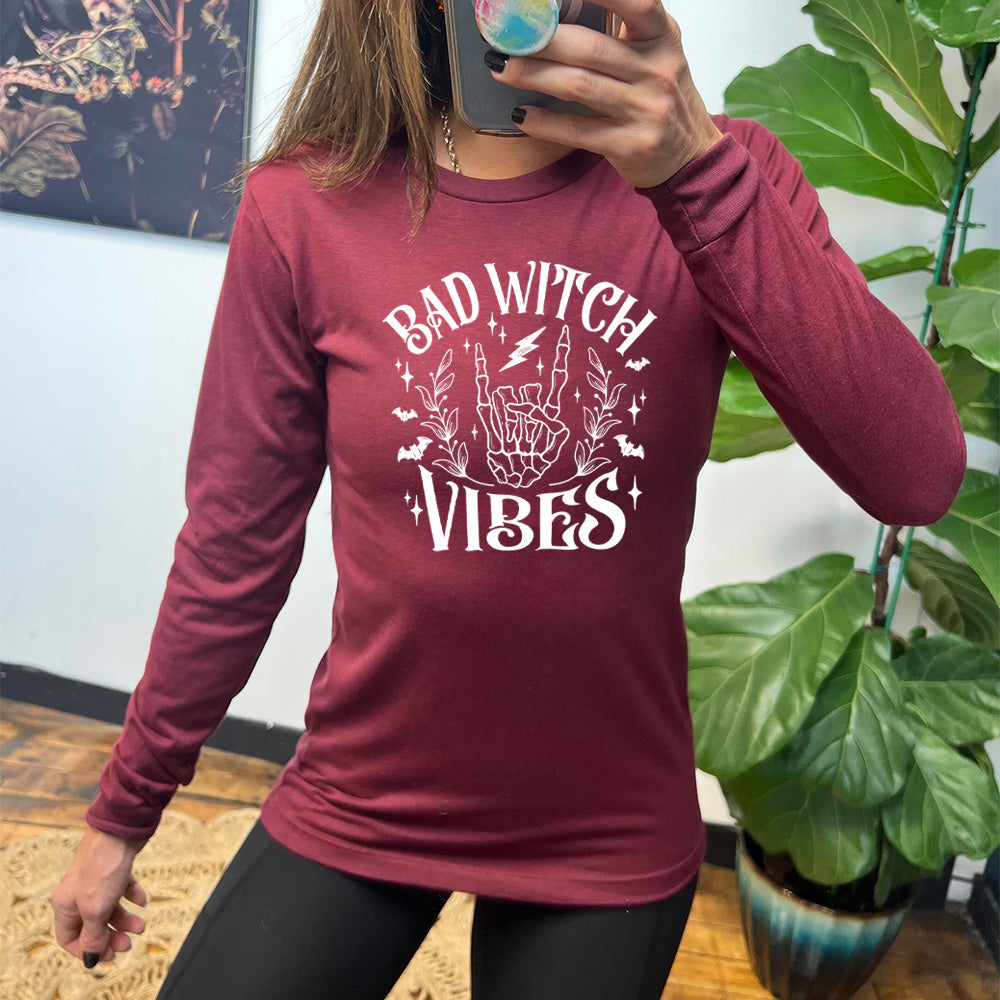 heather cardinal long sleeve shirt with the text "Bad Witch Vibes" on it, with the graphic of a skeletal rocker hand, a lightening bolt, some bats, a couple branches, and small stars