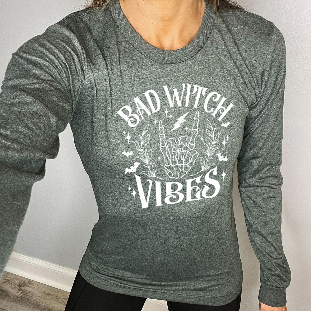 heather forest long sleeve shirt with the text "Bad Witch Vibes" on it, with the graphic of a skeletal rocker hand, a lightening bolt, some bats, a couple branches, and small stars