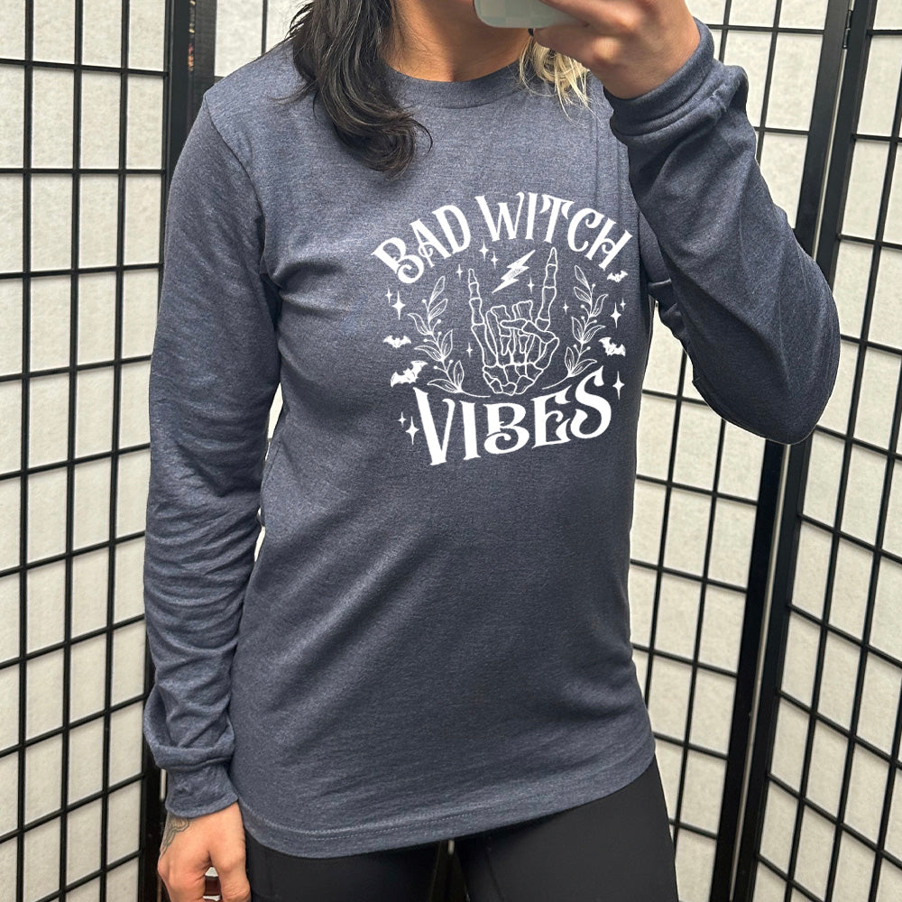 heather navy long sleeve shirt with the text "Bad Witch Vibes" on it, with the graphic of a skeletal rocker hand, a lightening bolt, some bats, a couple branches, and small stars