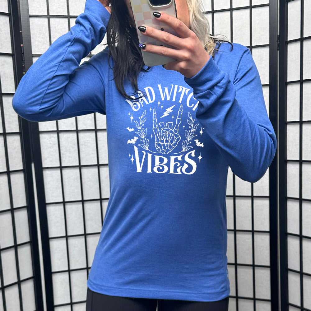 heather true royal long sleeve shirt with the text "Bad Witch Vibes" on it, with the graphic of a skeletal rocker hand, a lightening bolt, some bats, a couple branches, and small stars