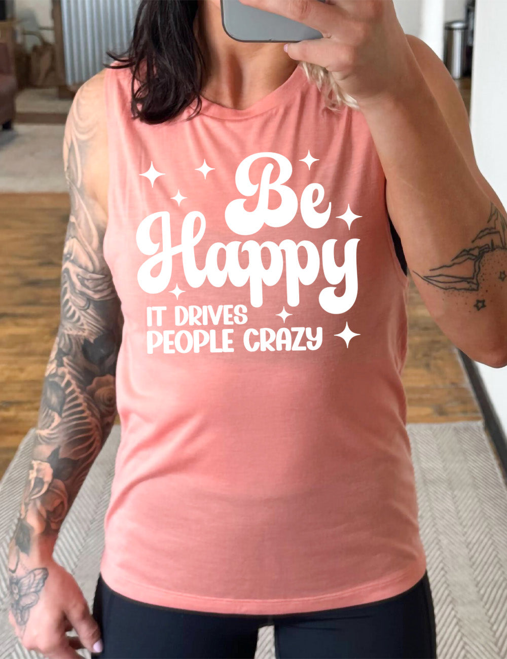 peach muscle tank with the words "be happy it drives people crazy" on it, with a number of stars around
