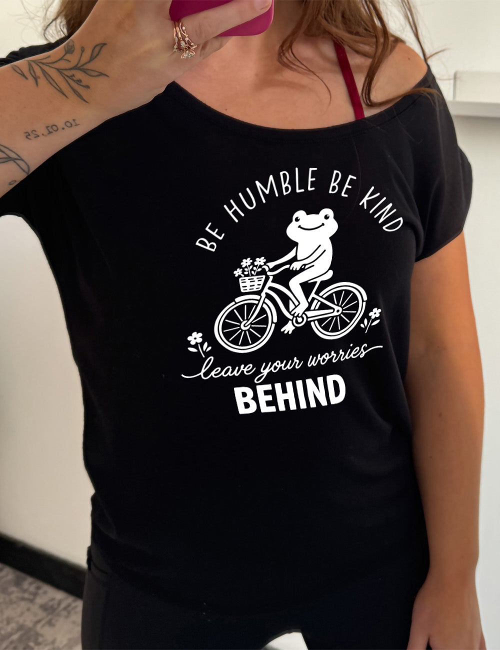 black slouchy tee with the graphic of a happy frog riding a bike that has a basket of flowers attached, with a flower in front and behind the bike, and the wording arching above and ending below saying "be humble be kind leave your worries BEHIND"