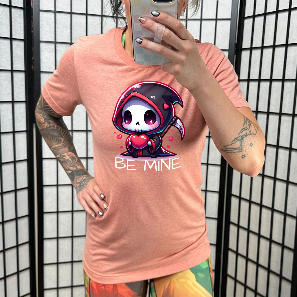 sunset unisex tee with large graphics of a cartoony grim reaper holding a heart, with little hearts surrounding, and the words "be mine" on it