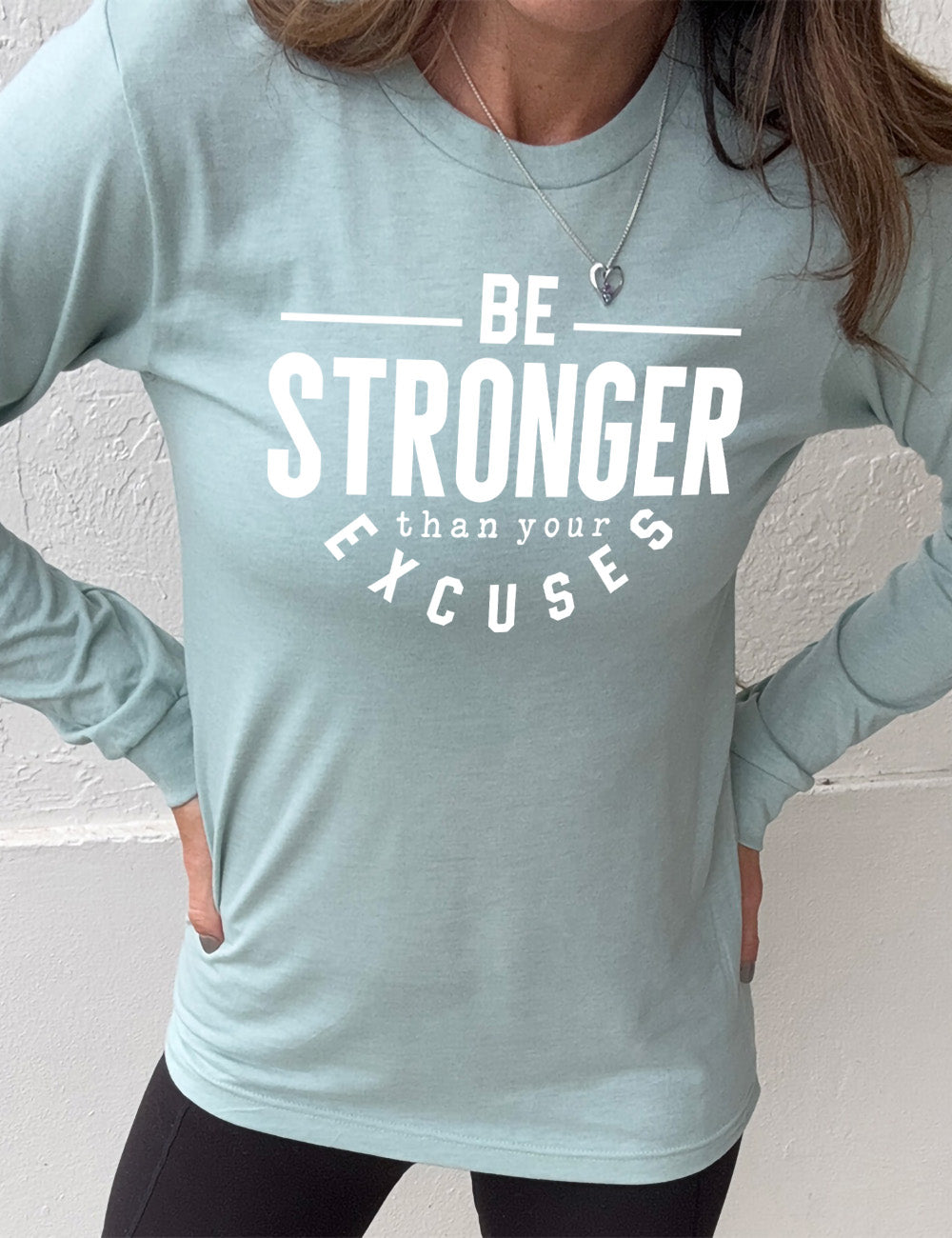 heather dusty blue long sleeve tee with words on it saying "be STRONGER than your excuses", with lines in front and after the word "be", the word "stronger" in the largest font, and the word "excuses" arching