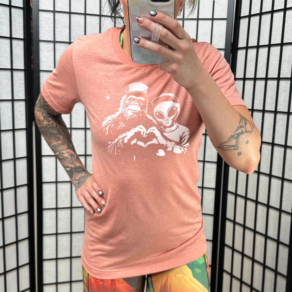Model wearing sunset colored shirt with bigfoot and alien doing heart hands graphic on it