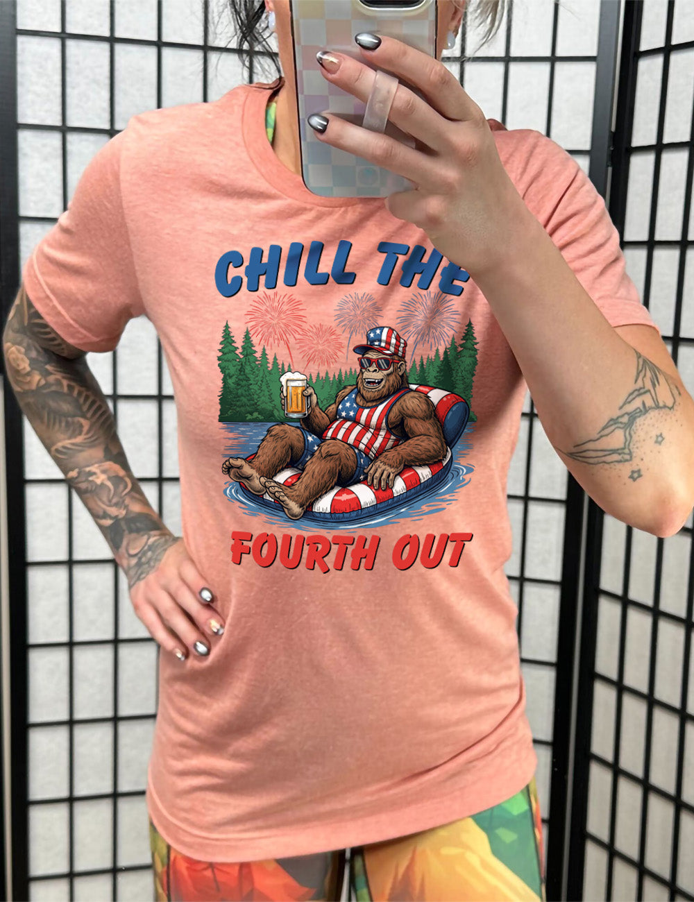 sunset unisex tee with the graphic of a grinning Bigfoot holding a beer, wearing sunglasses and an American flag outfit, in an American flag tube, in the water, with a background of trees, with fireworks above, and the words saying "chill the fourth out" on it