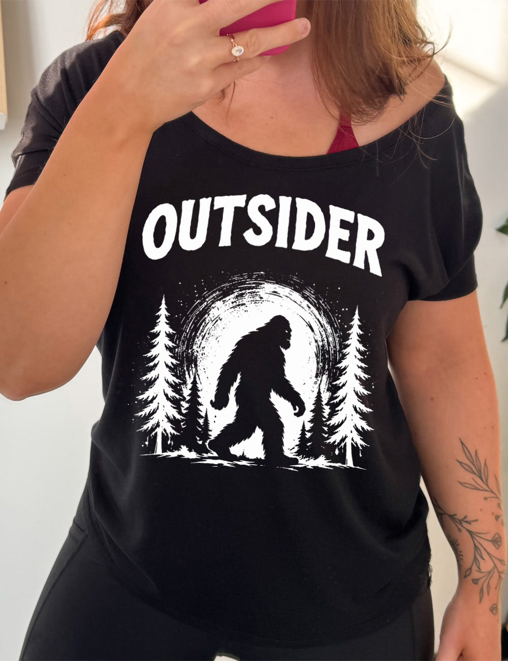 black slouchy tee with the graphic of bigfoot, silhouetted against a large moon, walking through pine trees, and a word above it saying "OUTSIDER"