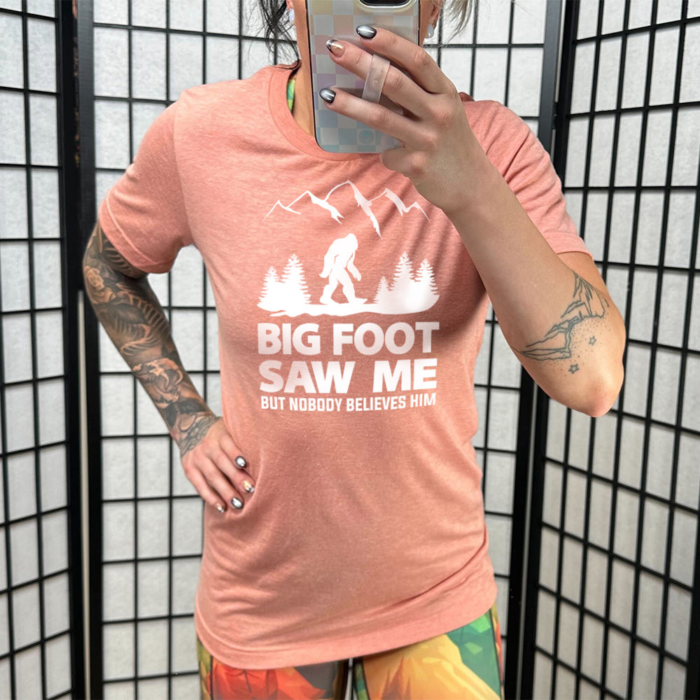 Model wearing sunset colored shirt with bigfoot in the woods graphic on it that says "Bigfoot Saw Me But Nobody Believes Him"