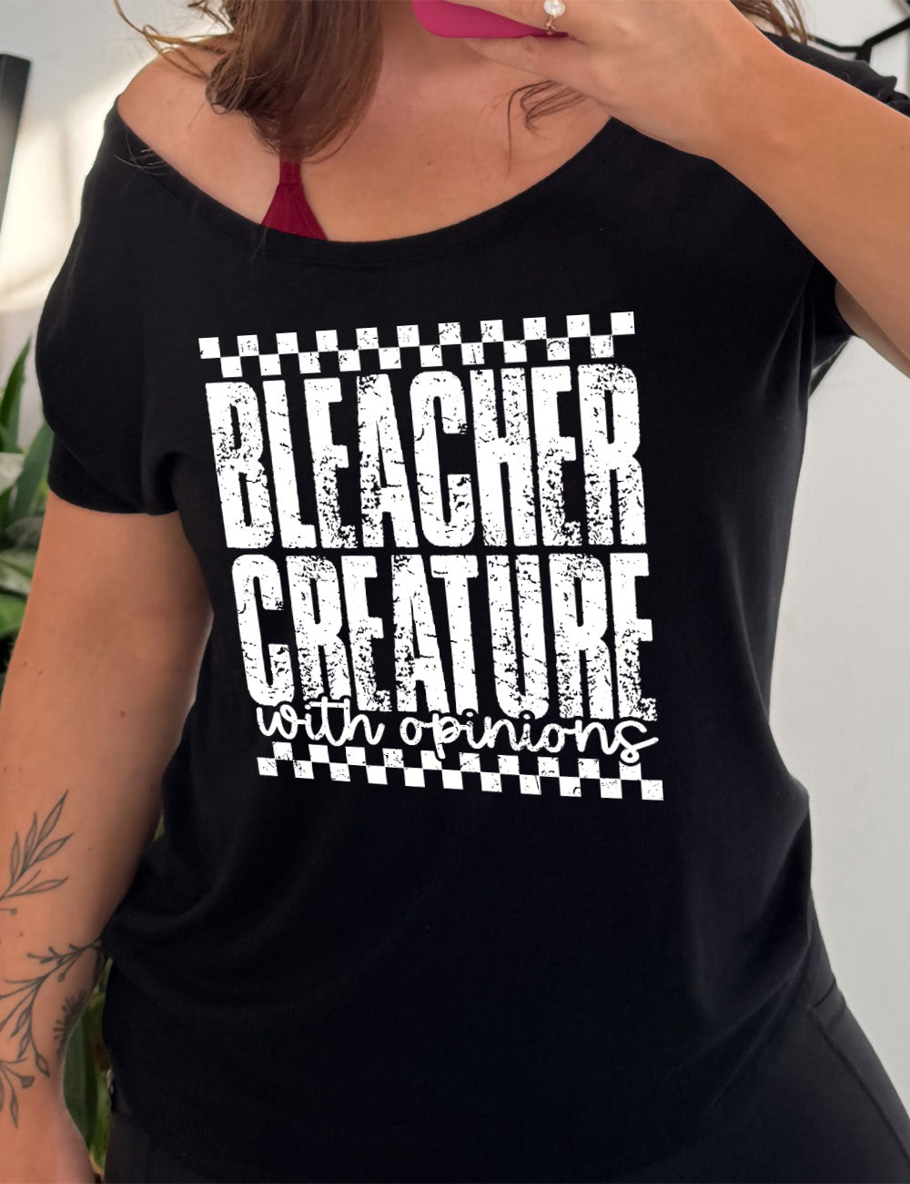 black slouchy tee with the large words "bleacher creature" and the small words underneath "with opinions", all of which has a top and bottom checkered border