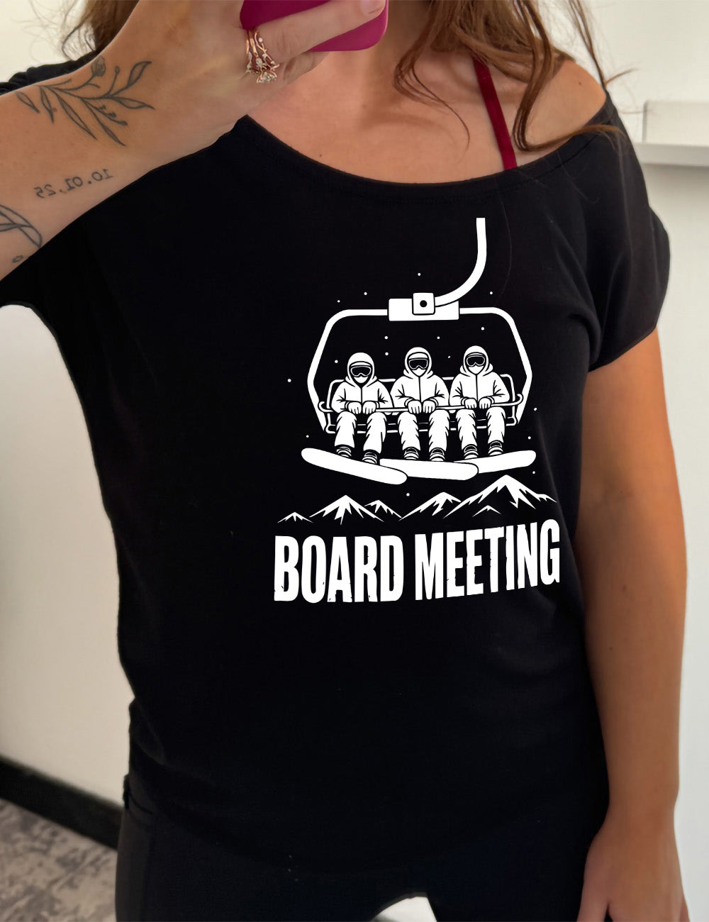 black slouchy tee with the graphic of three snowboarders riding a ski lift, with snowy mountains in the distance and little dots of snow around, and words underneath this saying "board meeting" in capital letters