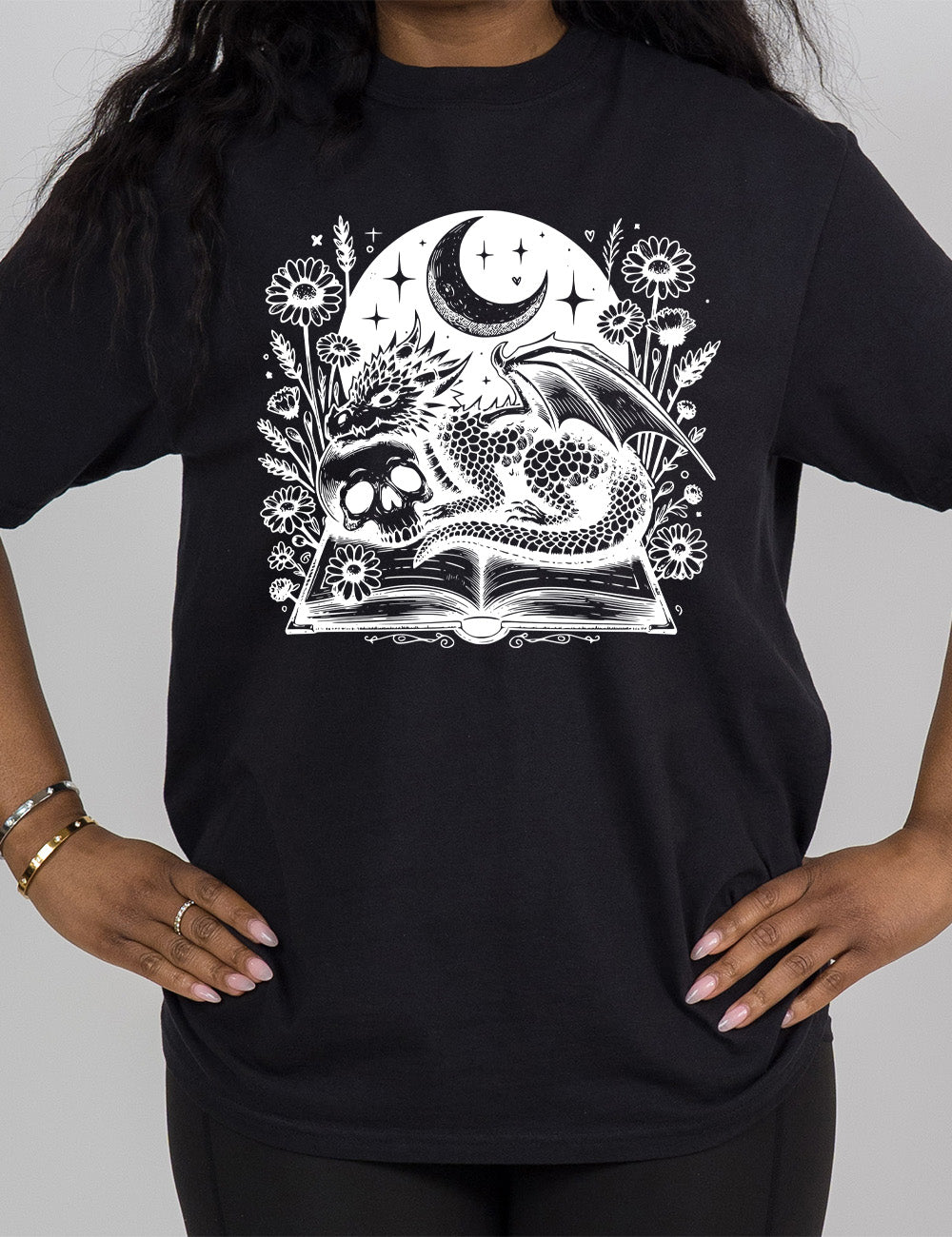 black modern tee with the very detailed and textured graphic of a dragon and a skull resting on an open book, with night sky behind it, with a large crescent moon, and many flowers growing around this scene