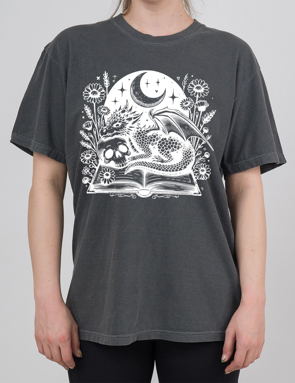 pepper modern tee with the very detailed and textured graphic of a dragon and a skull resting on an open book, with night sky behind it, with a large crescent moon, and many flowers growing around this scene