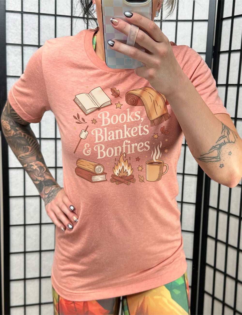sunset unisex tee with the words "books, blankets & bonfires" on it, surrounded by graphics including an open book, a scarf partially rolled up, a steaming beverage in a mug, a campfire, a book with a log on top of it, and a marshmallow on a stick, as well as a lot of little stars and leaves