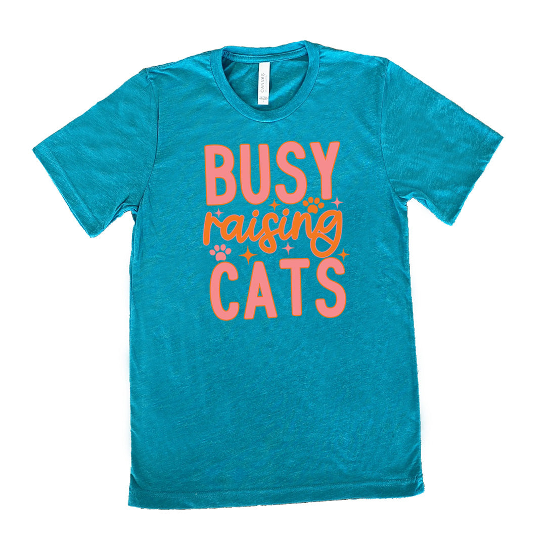 teal unisex tee with the words "busy raising cats" on it, with graphics of a couple small paw prints and a number of little stars