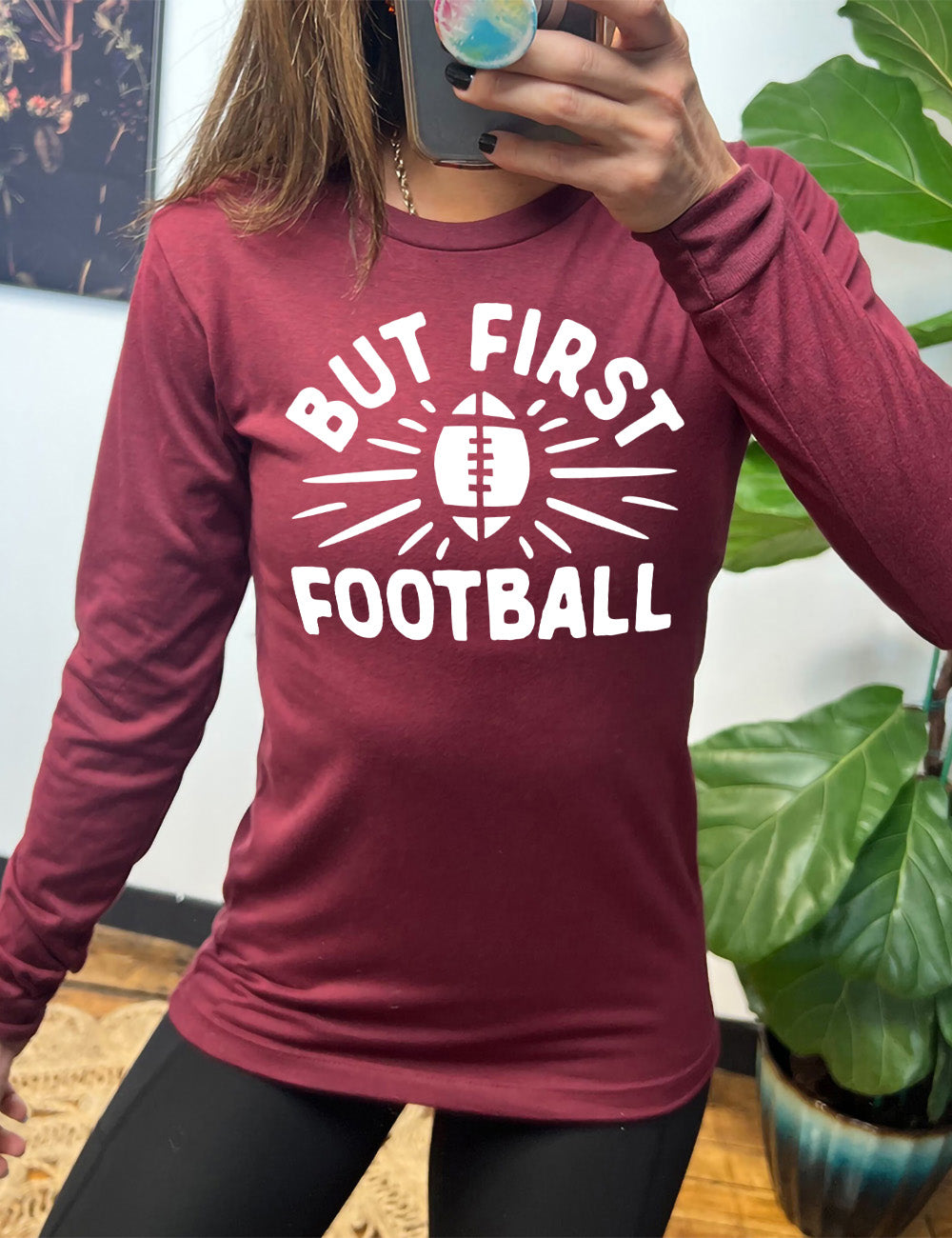 heather cardinal long sleeve with the words "but first football" in capital font, with "But first" arching over the graphic of a football, with rays beaming off of it