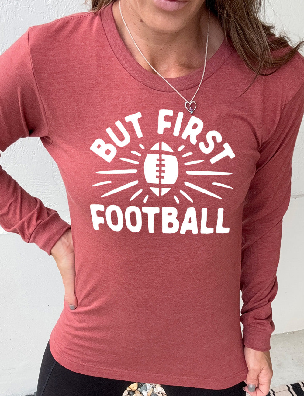 heather clay long sleeve with the words "but first football" in capital font, with "But first" arching over the graphic of a football, with rays beaming off of it