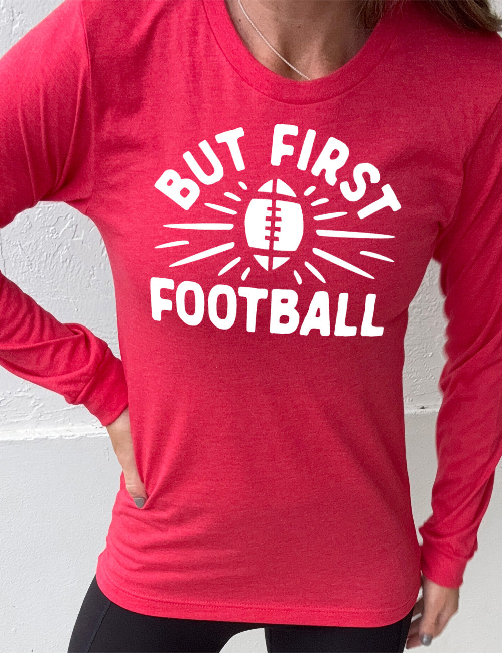 heather red long sleeve with the words "but first football" in capital font, with "But first" arching over the graphic of a football, with rays beaming off of it