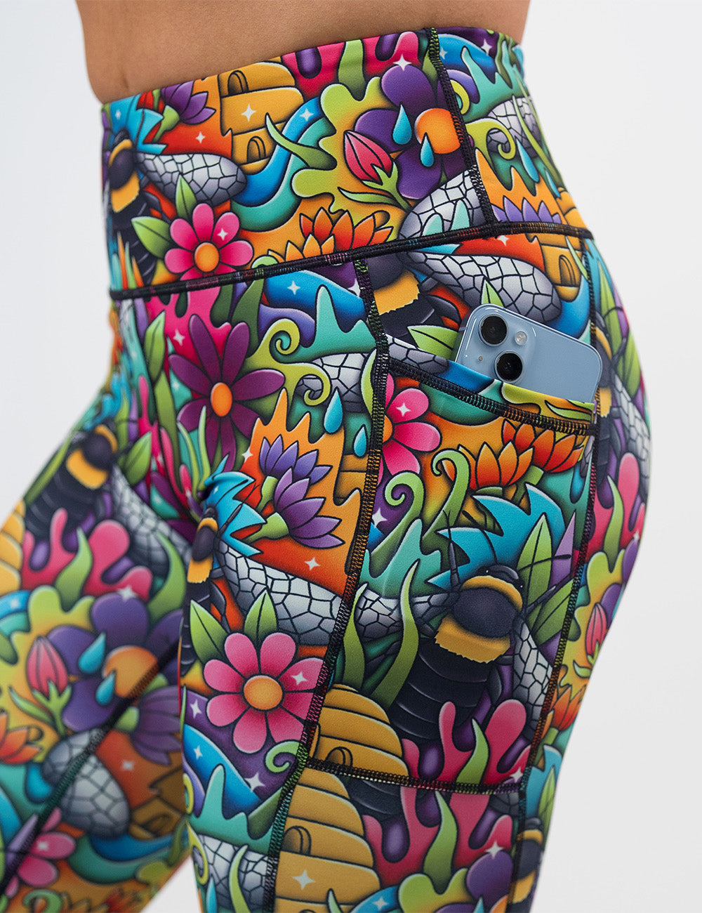 close up of the cartoon bees against bold colorful cartoon flowers pattern leggings pocket with a phone in it against a white background