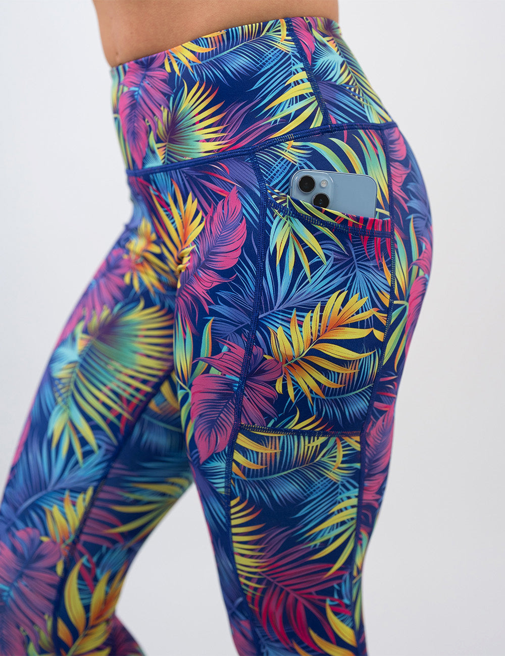 close up of a phone in the pocket of the vibrant rainforest leaves leggings against a white background