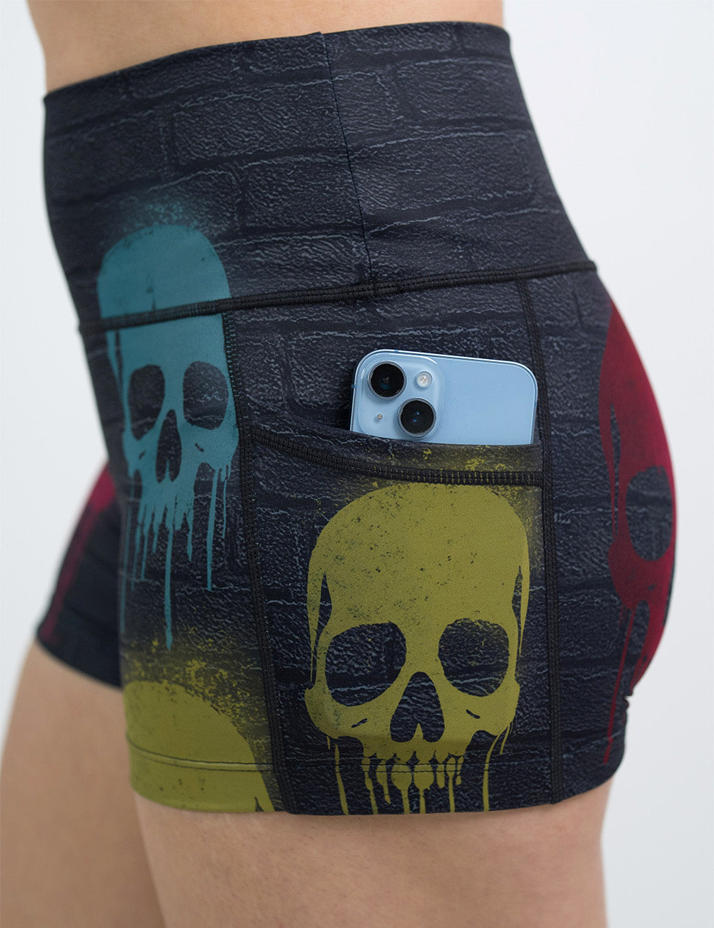 close up of the 2.5 inch black brick pattern with blue, red, and yellow skull spray paint patterns shorts with a phone in its pocket