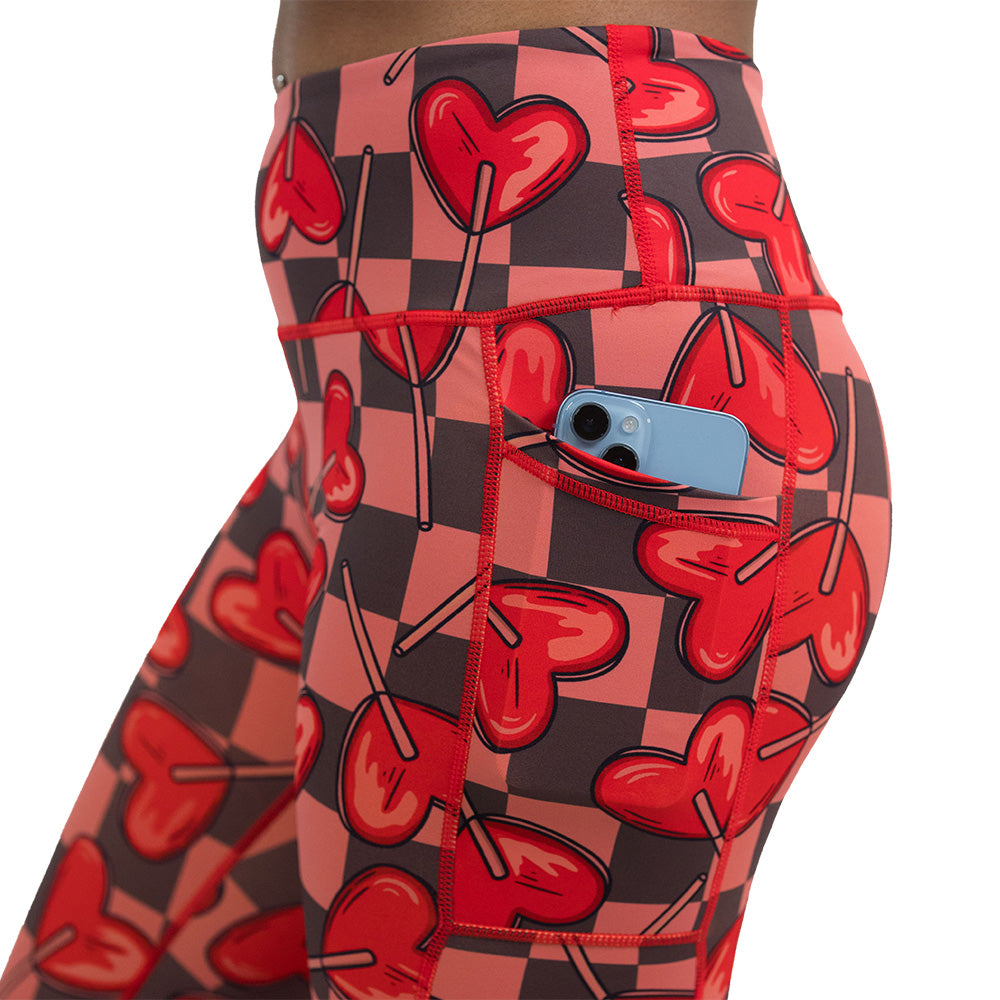 close up of the pocket of heart lollypops on a checker board background pattern leggings with a phone in the pocket against a white background