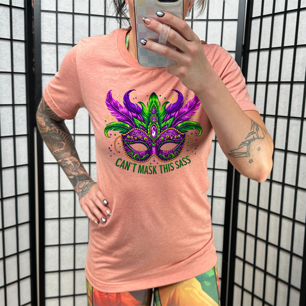 sunset unisex tee with the graphic of a fancy, feathered and bejeweled, and colorful mardi gras mask, and words underneath saying "can't mask this sass"
