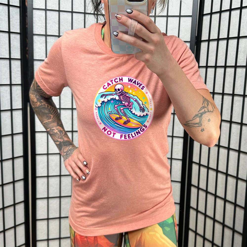 Model wearing sunset colored shirt with a skeleton surfing graphic on it that says "Catch Waves Not Feelings"