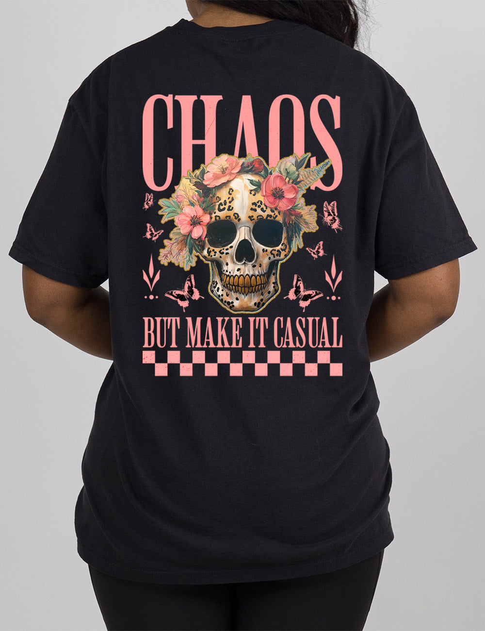 black modern tee with the print placement on the back, with the large word "chaos" on it, with the smaller words "but make it casual," and the graphic in between this of a gold-toothed skull with some leopard print, with a crown of flowers, surrounded by butterflies