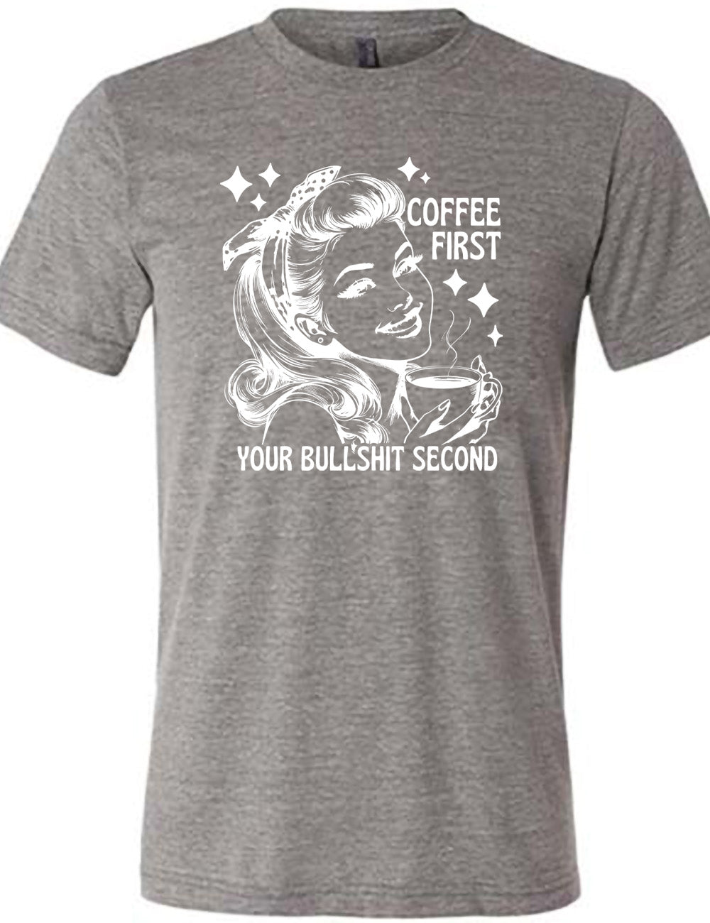 grey unisex tee with the graphic of a woman happily holding a mug of steaming coffee, with the words "coffee first" (next to her face) and "your bullshit second" underneath, and a few stars around her