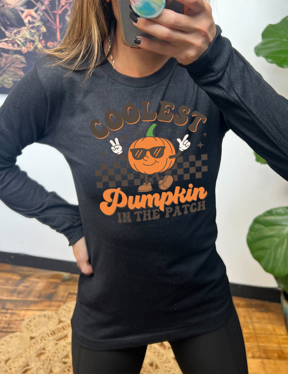 heather black long sleeve with the graphic of a pumpkin, wearing sunglasses, both arms raised, foot kicking out, in front of a checkered banner, surrounded by a few little stars, with the words around it saying "coolest pumpkin in the patch"