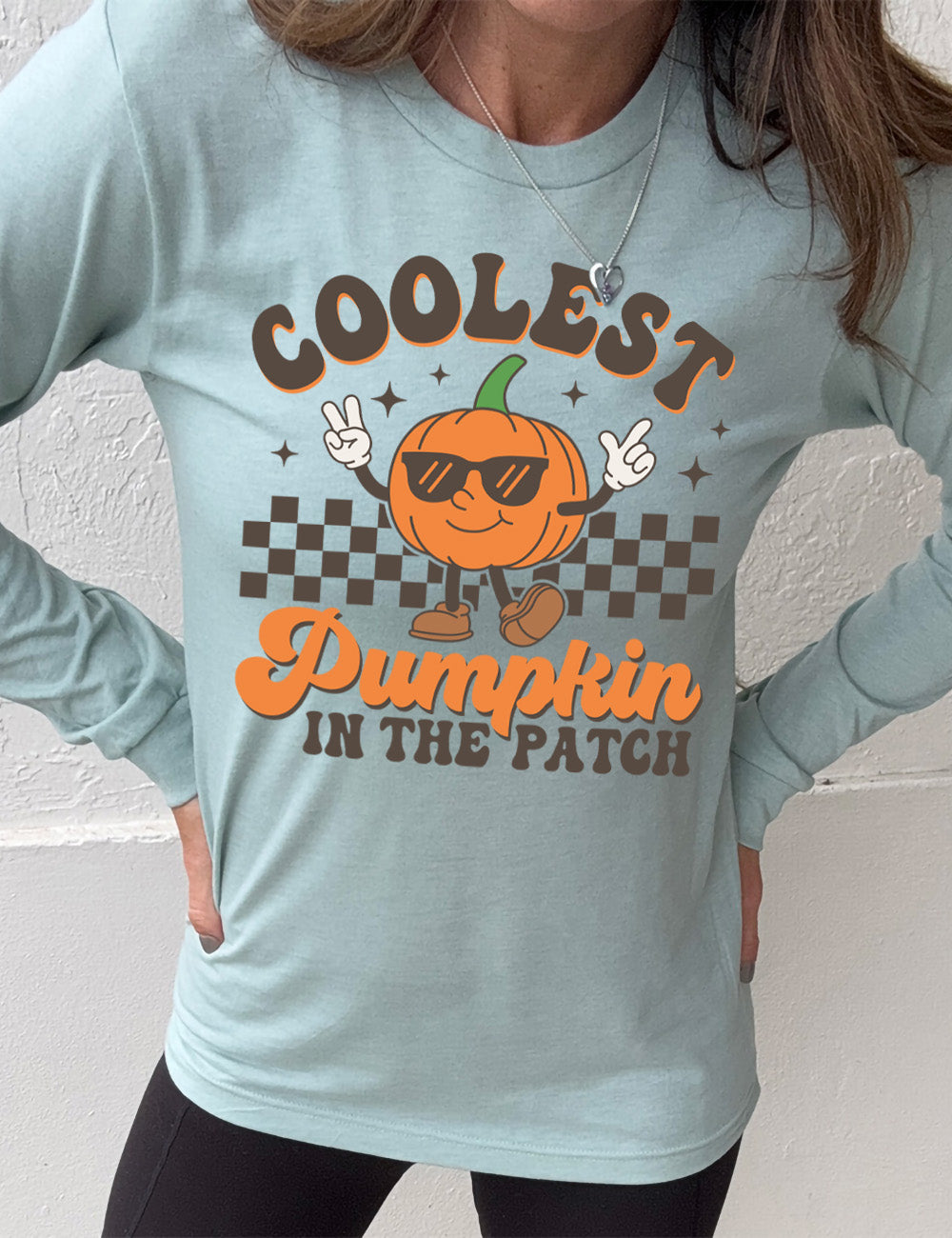 heather dusty blue long sleeve with the graphic of a pumpkin, wearing sunglasses, both arms raised, foot kicking out, in front of a checkered banner, surrounded by a few little stars, with the words around it saying "coolest pumpkin in the patch"