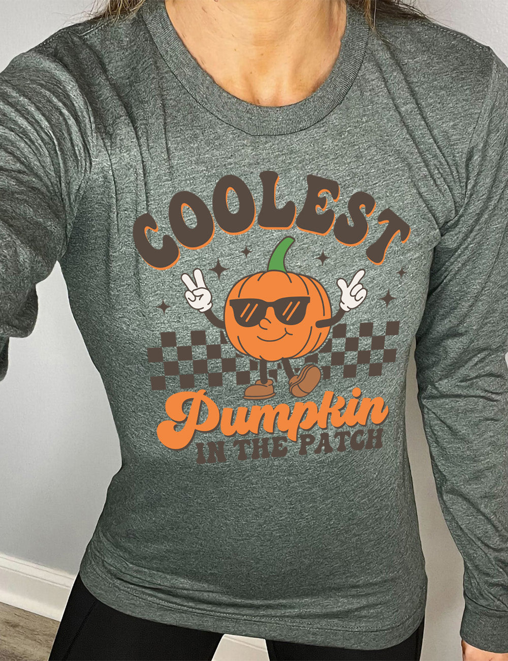 heather forest long sleeve with the graphic of a pumpkin, wearing sunglasses, both arms raised, foot kicking out, in front of a checkered banner, surrounded by a few little stars, with the words around it saying "coolest pumpkin in the patch"