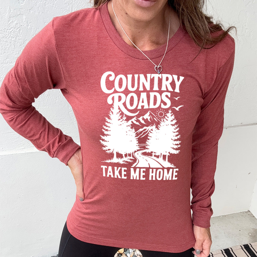 heather clay long sleeve with the words "country roads take me home" on it, and the graphic of a road going through tall trees, with mountains beyond, and the sun