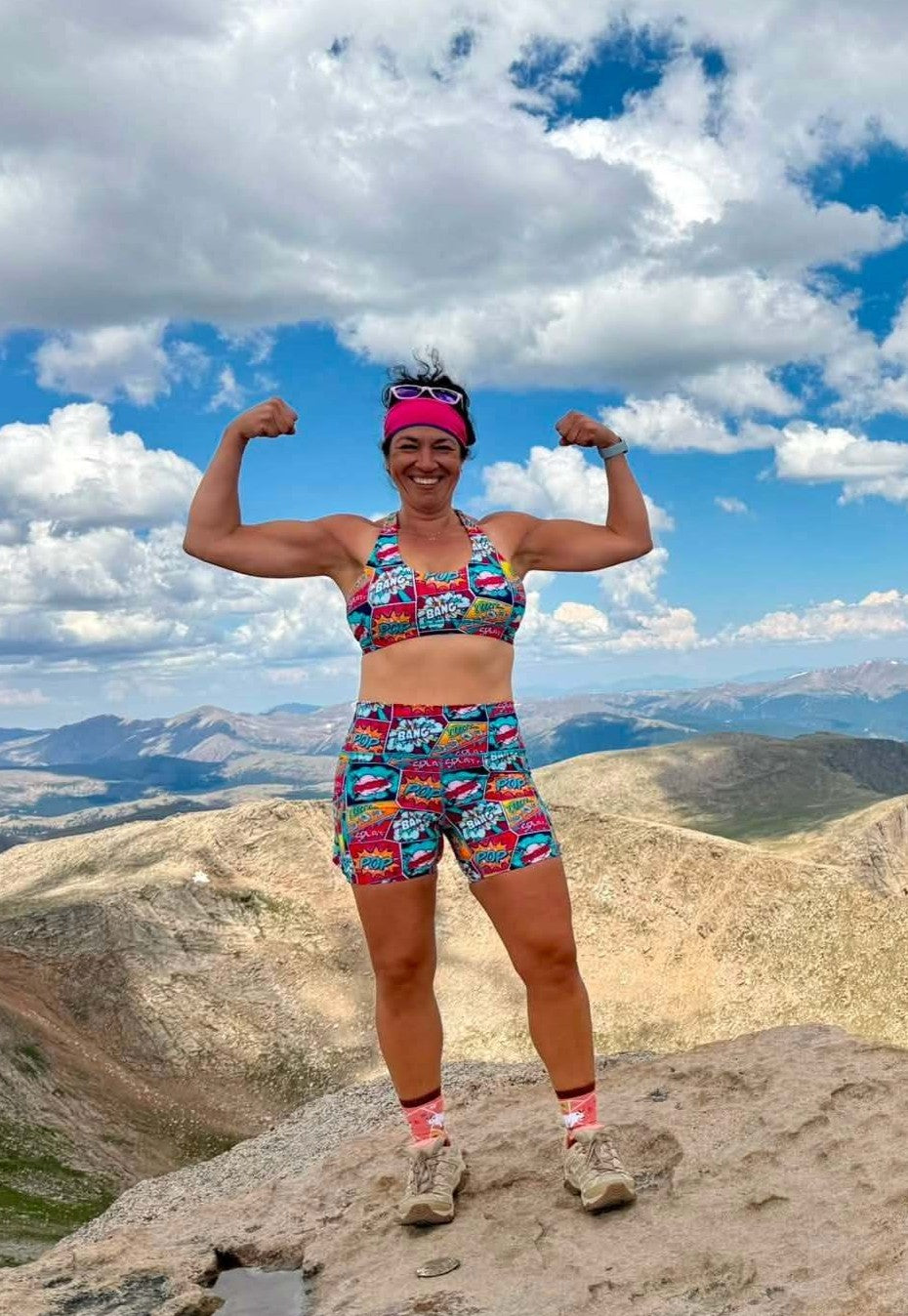 A woman posing on a mountain peak wearing a colorful, patterned athletic set consisting of a sports bra and matching shorts, with a pink visor and hiking shoes, against a backdrop of mountains and a cloudy blue sky.