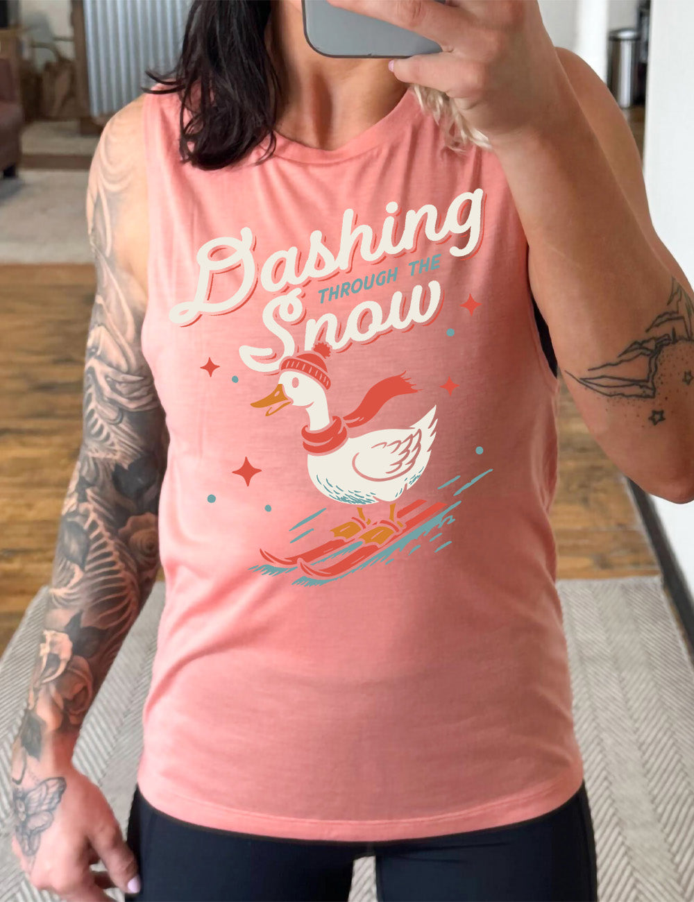 peach muscle tank with the graphic of a goose, wearing a hat and scarf, on skis, in downwards motion, with some stars, and the words above it saying "Dashing through the Snow"