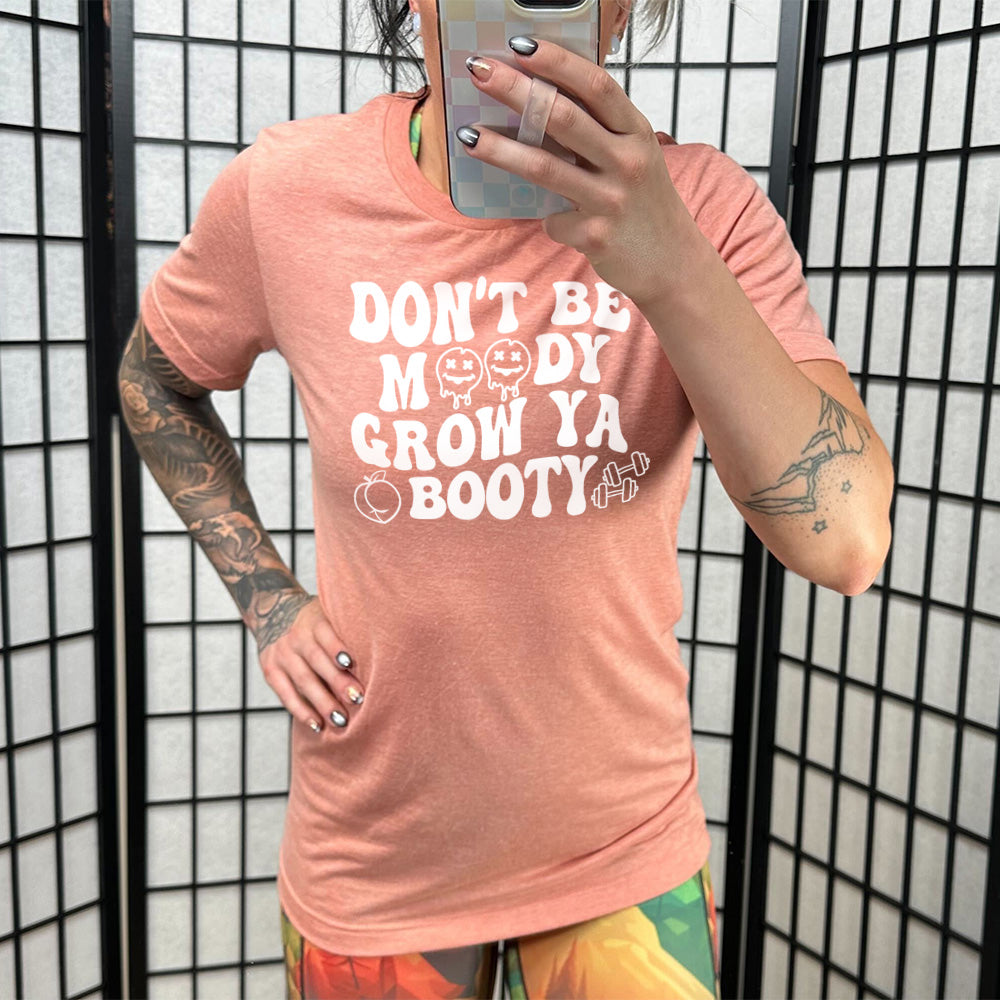 Sunset Unisex Tee with Don't Be Moody Grow Ya Booty in white lettering, the o's are emojis, and there's a peach and two barbells