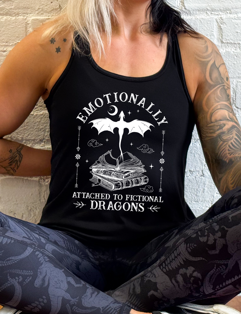 black racerback flowy tank with the words "emotionally attached to fictional dragons" on it, with graphics of a stack of books, with a dragon flying out of the open book on top, few a few little clouds, a moon, some stars, and arrows and flowers