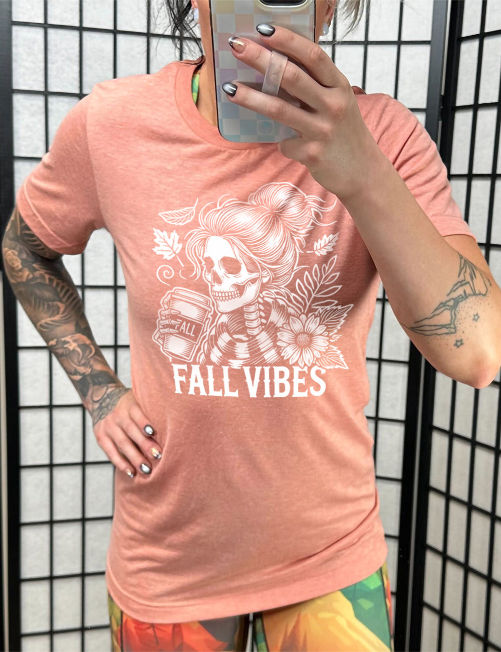 sunset unisex tee with the graphic of a skeletal woman, with her hair in a bun, holding a takeaway coffee cup labeled "fall", with a flower and leaves next to her, and the words underneath saying "fall vibes"