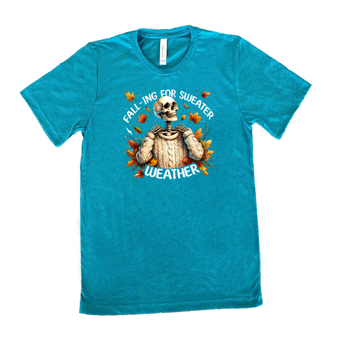 Teal colored shirt with fall leaves and skeleton wearing a sweater graphic on it that says "Fall-ing For Sweater Weather"