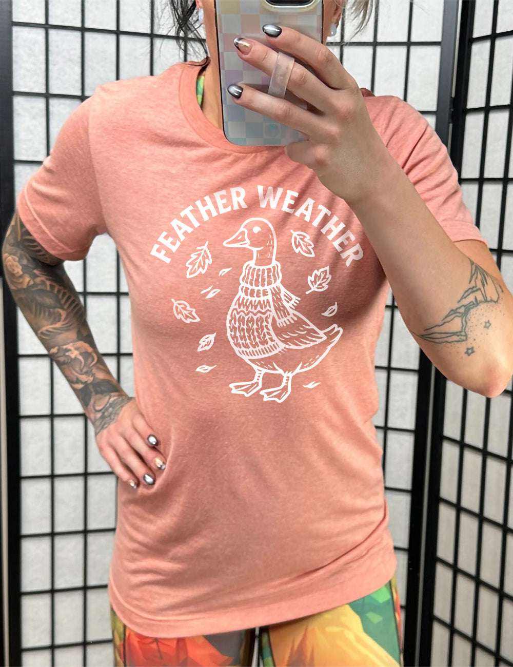 sunset unisex tee with the graphic of a goose, wearing a scarf and sweater, surrounded by falling leaves, and the words arched over it saying "feather weather"