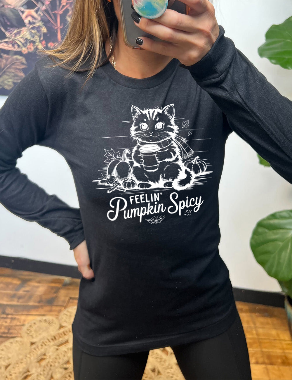 heather black long sleeve with the graphic of a cat, wearing a scarf, next to a few pumpkins of various sizes and a few falling leaves, holding a takeaway coffee, and the words underneath saying "feelin' pumpkin spicy"