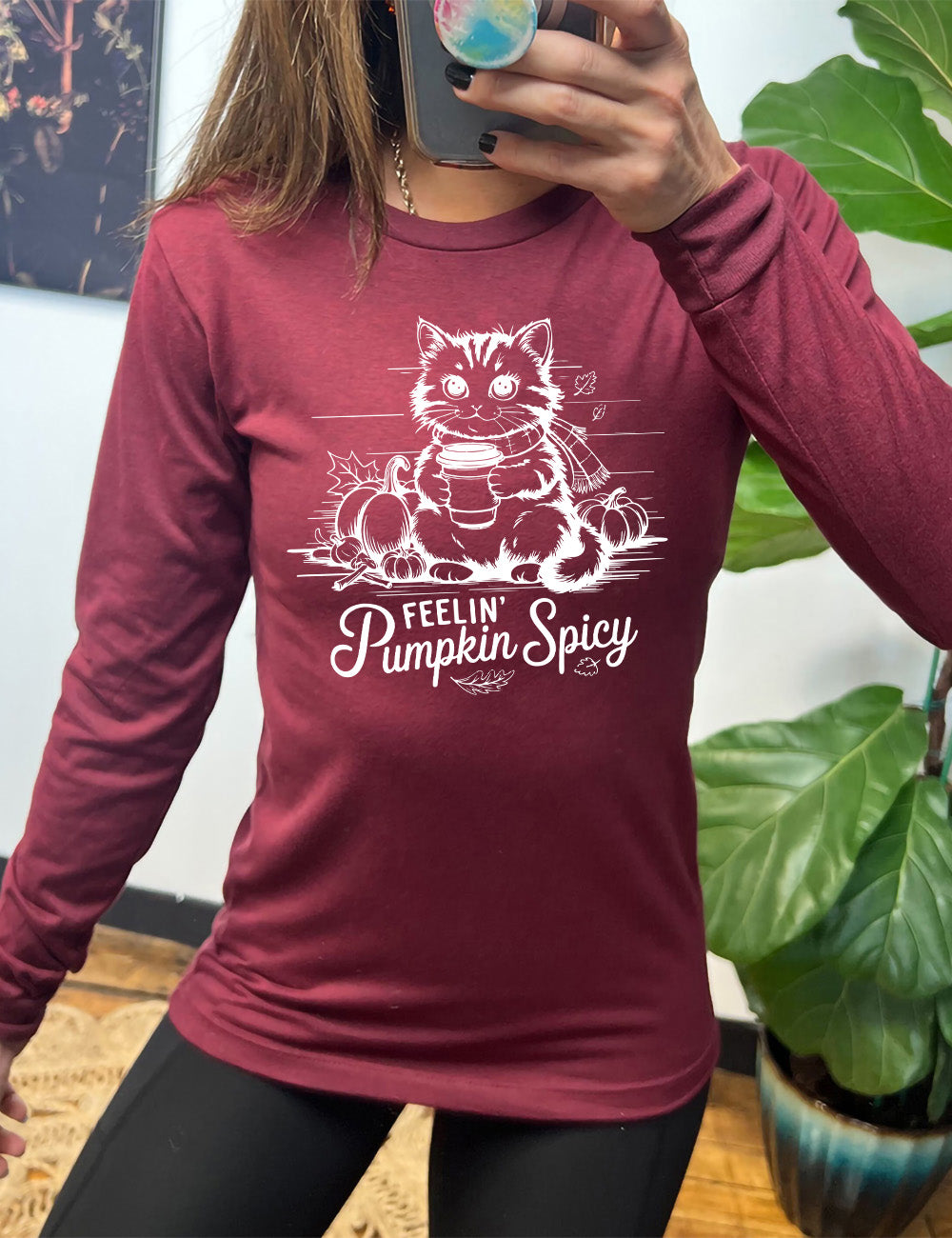 heather cardinal long sleeve with the graphic of a cat, wearing a scarf, next to a few pumpkins of various sizes and a few falling leaves, holding a takeaway coffee, and the words underneath saying "feelin' pumpkin spicy"