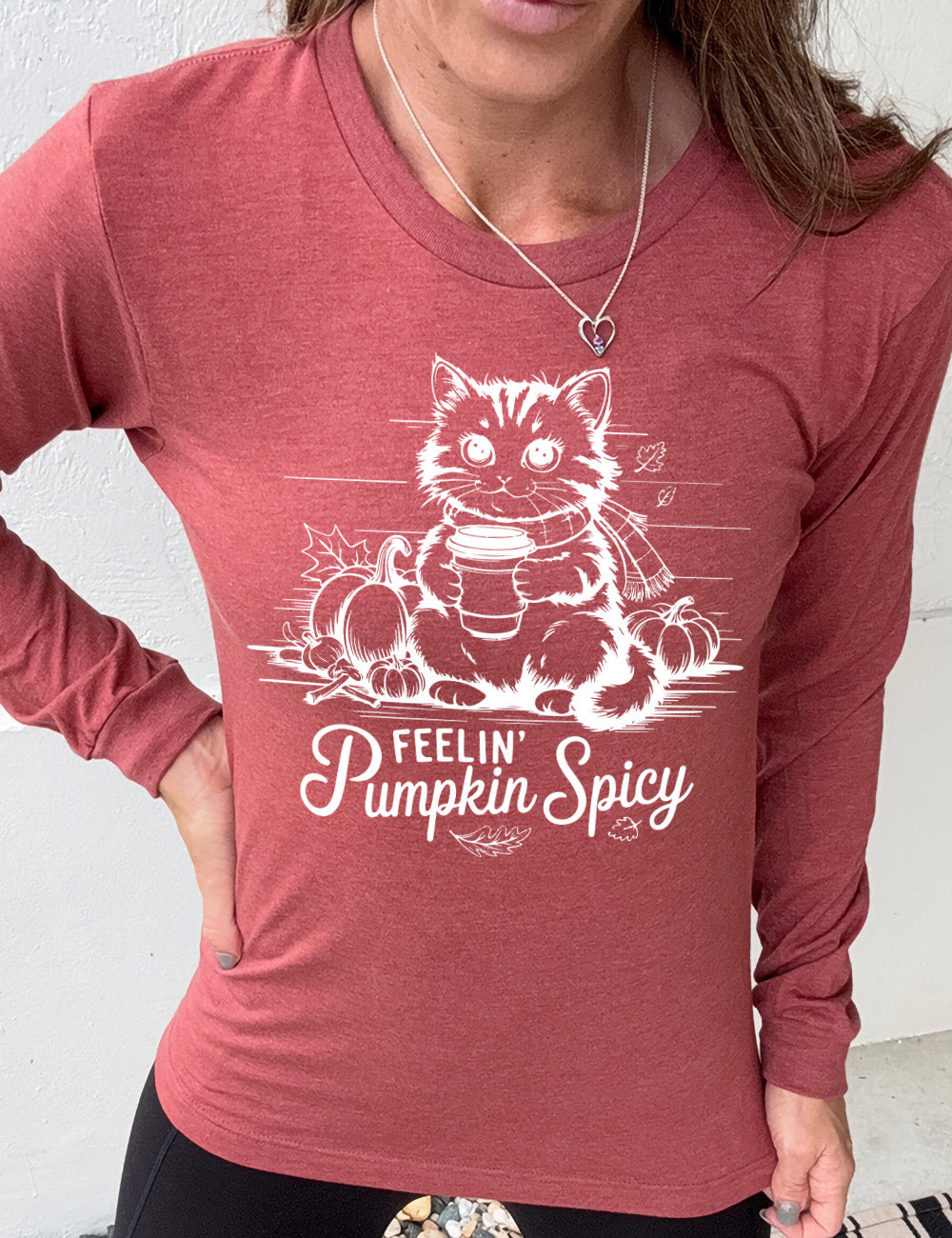 heather clay long sleeve with the graphic of a cat, wearing a scarf, next to a few pumpkins of various sizes and a few falling leaves, holding a takeaway coffee, and the words underneath saying "feelin' pumpkin spicy"