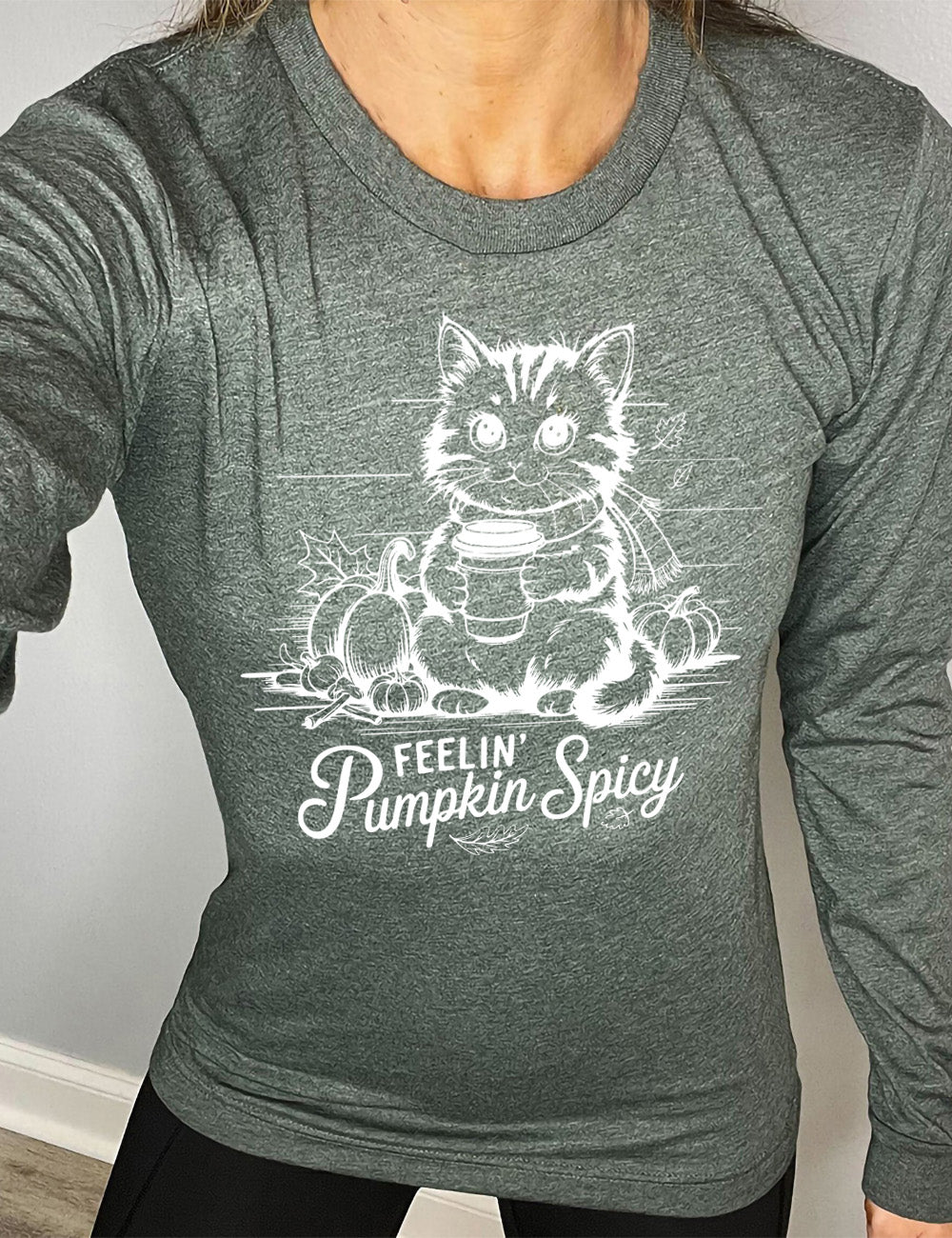 heather forest long sleeve with the graphic of a cat, wearing a scarf, next to a few pumpkins of various sizes and a few falling leaves, holding a takeaway coffee, and the words underneath saying "feelin' pumpkin spicy"
