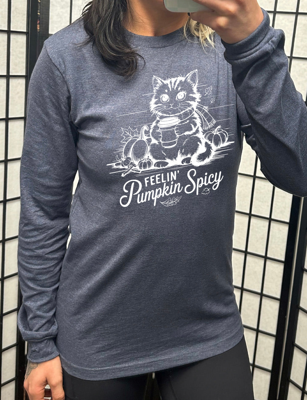 heather navy long sleeve with the graphic of a cat, wearing a scarf, next to a few pumpkins of various sizes and a few falling leaves, holding a takeaway coffee, and the words underneath saying "feelin' pumpkin spicy"